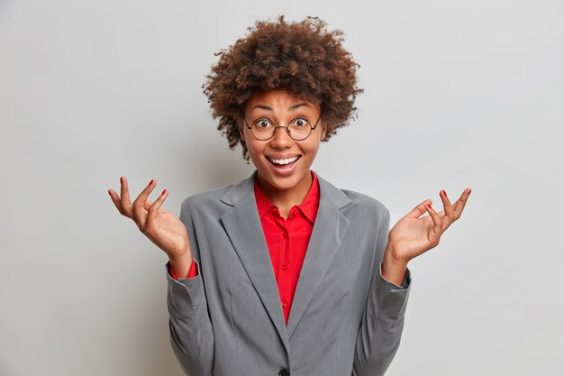 Gerente de oficina experto, de piel oscura, confuso y guapo, extiende las palmas de las manos en un gesto desorientado, tiene una expresión feliz, es interrogado, usa una camisa roja y una elegante chaqueta gris