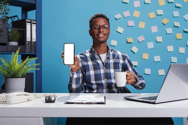 Gerente de proyecto apuntando dispositivo de teléfono con pantalla de maqueta a cámara. Empresario adulto joven sentado en la oficina del espacio de trabajo moderno de la empresa mientras muestra una plantilla aislada de pantalla blanca a la cámara.