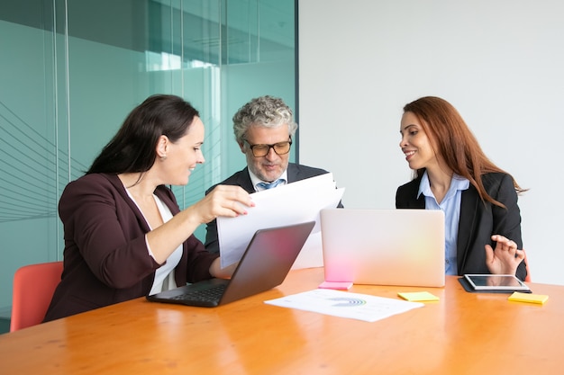 Los gerentes presentan informes en papel al jefe. Hombre de pelo gris en traje y dos mujeres de negocios revisando papeles juntos.