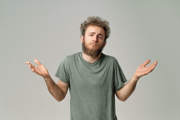 Gesticular NO SÉ o LO SIENTO joven apuesto hombre de pelo rizado salvaje con barba con ojos azules brillantes aislado sobre fondo gris Hombre pensante joven en camiseta verde sobre blanco Copiar espacio