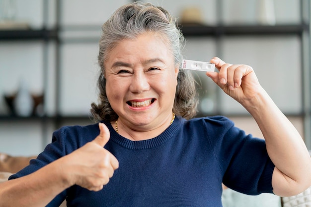 Gesto de retención de la mano de una anciana asiática jubilada y muestra el resultado del kit de prueba rápida covid19 con signo negativo con felicidad y prueba rápida de prueba alegre en casa