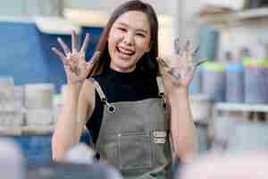 Foto gratuita gesto sonriente saludando a la artista crramista asiática mirando a la cámara artista alegre mirando a la cámara portriat mientras esculpe su nuevo trabajo en el taller de cerámica estudio de arte y creación femenina
