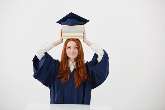 Ginger mujer graduada en manto sonriendo sosteniendo libros en la cabeza debajo de la tapa.