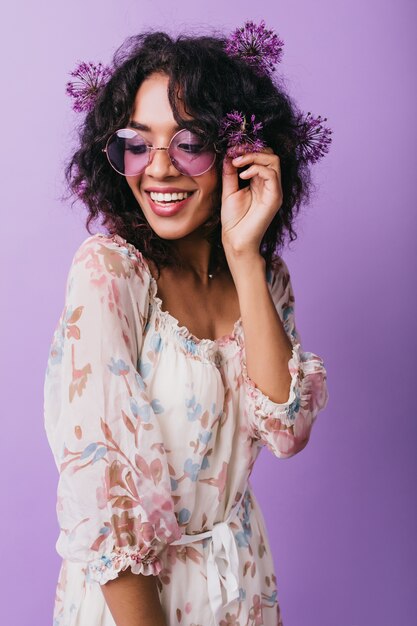 Glamorosa chica africana con gafas divirtiéndose durante la sesión de fotos con alliums. Foto interior de modelo femenino positivo vestido expresando felicidad.