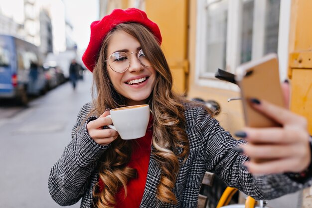 Glamorosa chica pálida en vasos haciendo selfie mientras bebe café en la cafetería al aire libre
