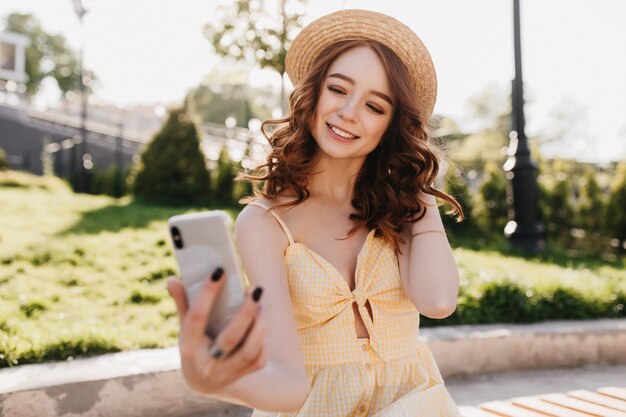 Glamorosa chica pelirroja con teléfono para selfie. Disparo al aire libre de la impresionante dama elegante en traje amarillo escalofriante en el parque.