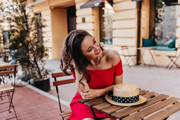 Glamorosa chica sentada en un café al aire libre con una sonrisa. Mujer morena atractiva en vestido rojo escalofriante en restaurante de la calle.