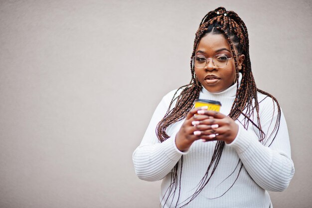 Glamorosa mujer afroamericana en suéter de cuello alto blanco posan en la calle con una taza de café a mano