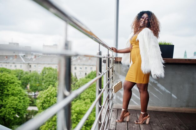 Glamour mujer afroamericana en vestido amarillo y capa de lana blanca con bolso posado en el balcón