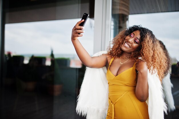 Glamour mujer afroamericana en vestido amarillo y capa de lana blanca haciendo selfie en teléfono