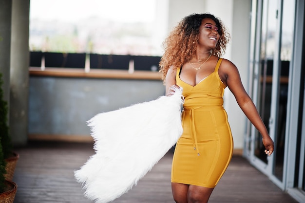 Glamour mujer afroamericana en vestido amarillo y capa de lana blanca posada en la terraza