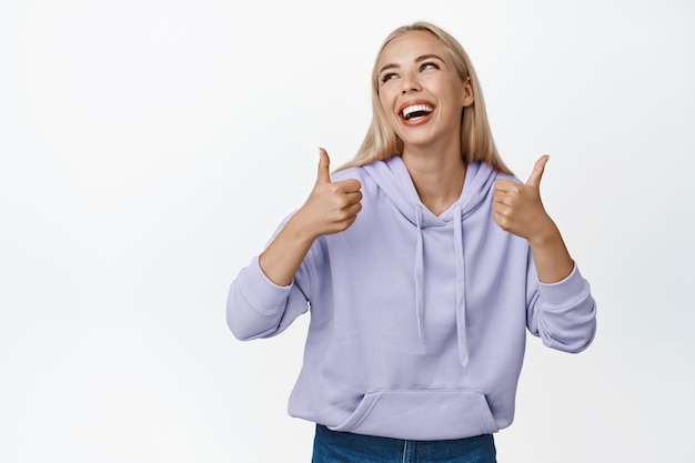 Glamour mujer rubia riendo sonriendo y mostrando los pulgares hacia arriba de pie feliz contra el fondo blanco.