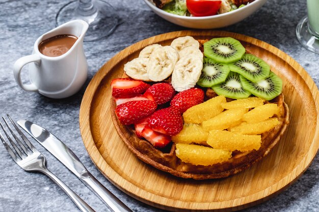 Gofre de vista lateral con kiwi, plátano, fresa, naranja y chocolate sobre la mesa