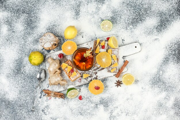 Gofres planos y obleas de arroz sobre una tabla de cortar blanca con té de hierbas, frutas cítricas, canela y colador de té sobre una superficie de mármol gris oscuro. horizontal