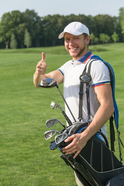 Foto gratuita golfista que muestra la señal aceptable y que sonríe para la cámara. hombre con sombrero de camionero blanco dando pulgar hacia arriba y sosteniendo una bolsa con conductores de golf.