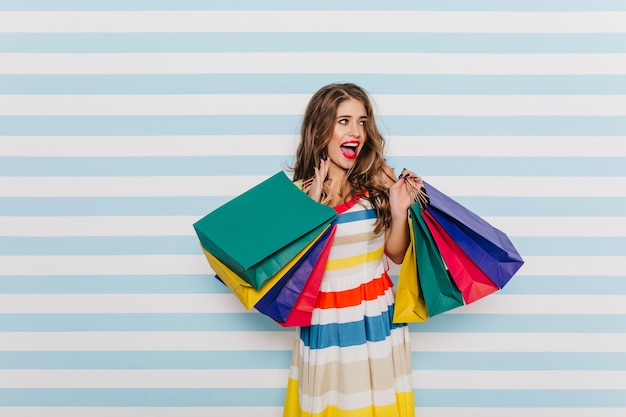 Gorgeos, elegante, joven mujer posando con coloridas bolsas de la compra. Señora de ojos azules de pelo oscuro con sonrisa feliz en la pared de luz