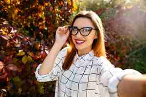 Foto gratuita gougeus modelo toma selfie mientras sostiene sus gafas con una mano en el jardín de otoño.