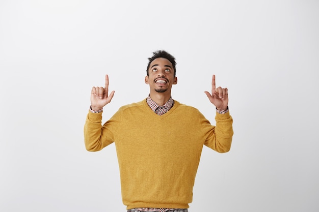 Gracias a dios es viernes. retrato de joven estudiante afroamericano interesado complacido en elegante suéter amarillo levantando las manos, mirando y apuntando hacia arriba, disfrutando de una bonita vista del cielo