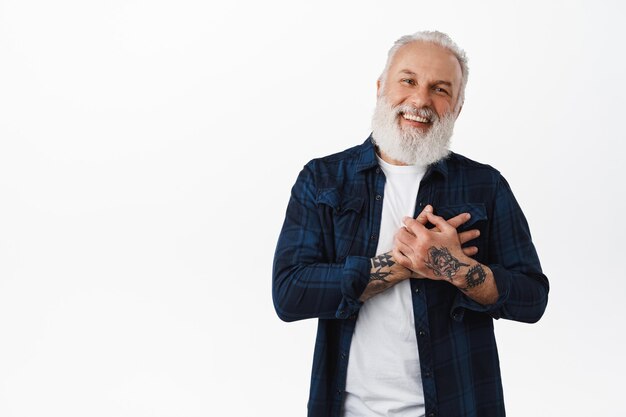 Gracias. Feliz hombre tatuado senior, tomados de la mano en el corazón y sonriendo, siendo halagado o tocado, luciendo sincero, complacido por algo, de pie sobre fondo blanco.