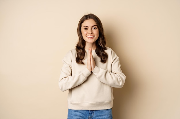 Gracias. Sonriente mujer feliz mostrando oración, gesto de namaste, agradeciendo, complacida y agradecida sobre fondo beige.