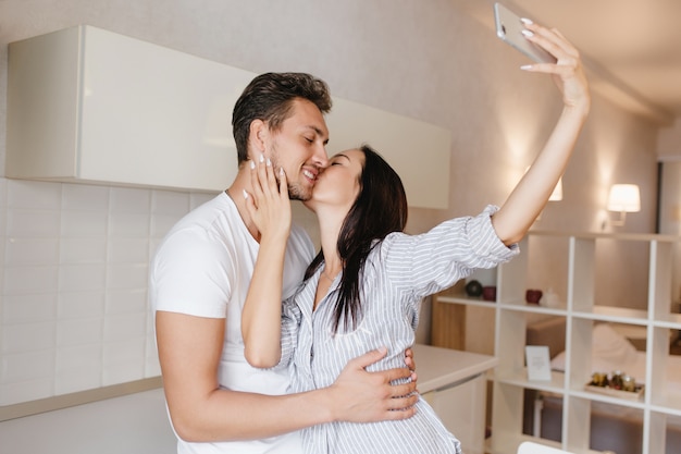 Graciosa dama de pelo negro en elegante camisa masculina haciendo selfie y besando a novio