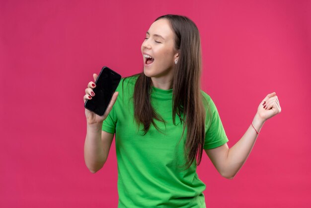 Graciosa hermosa joven vestida con camiseta verde sosteniendo smartphone usándolo como micrófono cantando sobre espacio rosa aislado