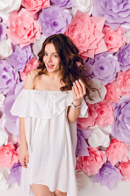 Graciosa linda chica morena con vestido blanco fuera del hombro se encuentra en un estudio con fondo de flores rosadas.