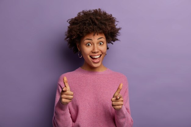 Graciosa mujer afroamericana feliz con peinado rizado apunta con pistolas a la cámara, finge disparar, te elige o te escoge, dice bang bang