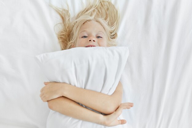 Graciosa niña con cabello rubio y ojos azules, divirtiéndose en la cama, abrazando la almohada blanca, va a conciliar el sueño. Niño feliz con almohada en casa, relajarse en el dormitorio. Estilo de vida de los niños