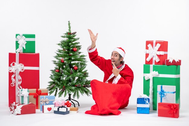 Gracioso adulto joven emocionado vestido como Papá Noel con regalos y árbol de Navidad decorado sentado en el suelo apuntando arriba sobre fondo blanco.