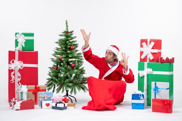 Gracioso adulto joven vestido como Papá Noel con regalos y árbol de Navidad decorado sentado en el suelo apuntando hacia arriba haciendo gesto de anteojos sobre fondo blanco.