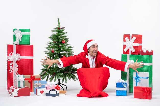 Gracioso feliz joven vestido como Papá Noel con regalos y árbol de Navidad decorado sentado en el suelo sobre fondo blanco.