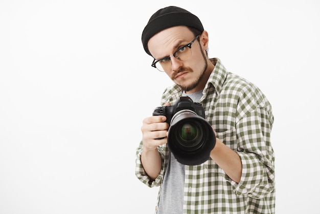 Foto gratuita gracioso fotógrafo masculino de aspecto serio con gorro negro y camisa a cuadros apuntando con una cámara profesional hacia adelante y mirando seriamente para tomar una foto durante el trabajo