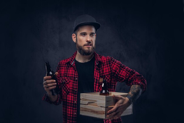 Gracioso hipster borracho barbudo sostiene una caja de madera con botellas de cerveza, de pie en el estudio.