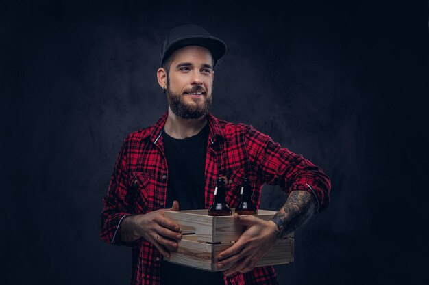 Gracioso hipster borracho barbudo sostiene una caja de madera con botellas de cerveza, de pie en el estudio.