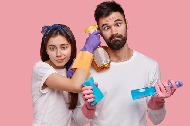 Gracioso hombre sin afeitar cruza los ojos, ayuda a la esposa a limpiar la casa, trabajar juntos, hacer las tareas domésticas, usar detergentes, esponja