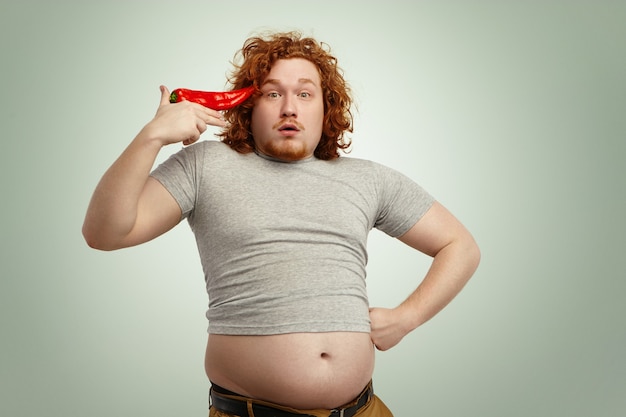 Gracioso hombre gordo con sobrepeso con cabello rizado de jengibre sosteniendo gran chile en su sien como una pistola