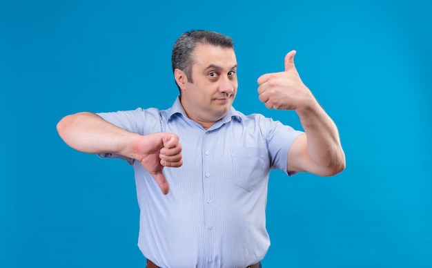 Gracioso hombre de mediana edad en camisa a rayas azul mostrando los pulgares hacia arriba y hacia abajo en un espacio azul