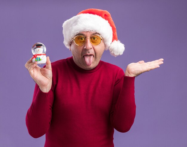 Gracioso hombre de mediana edad vestido con gorro de Papá Noel de Navidad en gafas amarillas sosteniendo bola de nieve de Navidad sacando la lengua con el brazo levantado sobre fondo púrpura