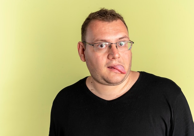 Gracioso hombre con sobrepeso en gafas con camiseta negra sacando la lengua sobre la luz