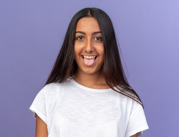 Gracioso joven en camiseta blanca mirando a la cámara con cara feliz sacando la lengua de pie sobre fondo azul.