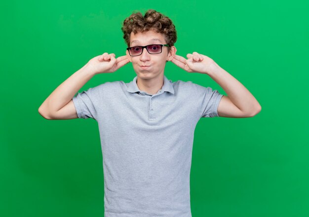 Gracioso joven de gafas negras con camisa de polo gris mostrando sus orejas mejillas blowiong feliz y alegre de pie sobre la pared verde