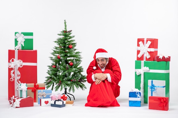 Gracioso joven vestido como Papá Noel con regalos y árbol de Navidad decorado sentado en el suelo sobre fondo blanco.