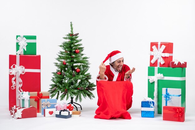 Gracioso joven vestido como Papá Noel con regalos y árbol de Navidad decorado sobre fondo blanco.