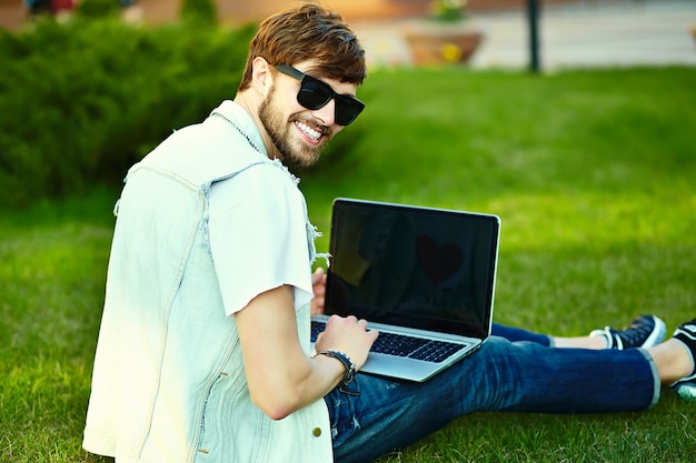 Gracioso sonriente hipster hombre guapo en elegante ropa de verano en la calle sentado en el césped en el parque con notebook