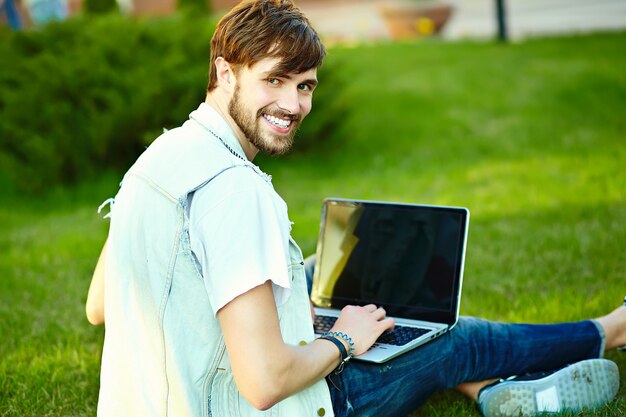 Gracioso sonriente hipster hombre guapo en elegante tela de verano en pasto con notebook