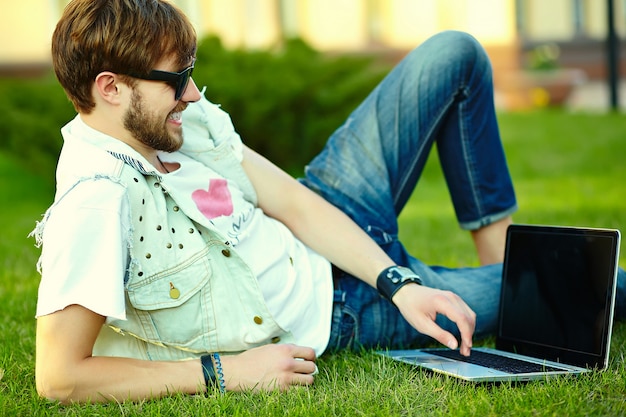 Gracioso sonriente hipster hombre guapo en elegante tela de verano en pasto con notebook