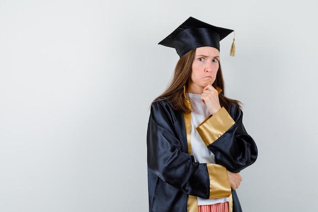 Graduada femenina apoyando la barbilla en la mano con vestimenta académica y mirando pensativa, vista frontal.