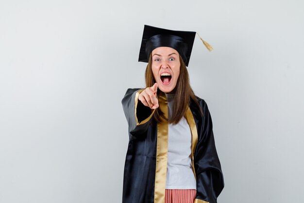 Graduada femenina apuntando a la cámara en traje académico y mirando agresivo, vista frontal.