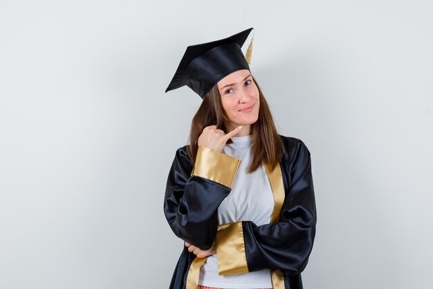 Graduada femenina apuntando a la esquina superior derecha en uniforme, ropa casual y luciendo esperanzada, vista frontal.